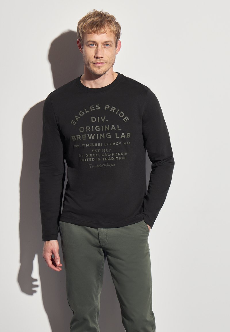 Musta pitkähihaine paita, jossa on tummanharmaa tekstuurikuvio, yhdistettynä vaaleanvihreisiin housuihin, esillä neutraalia taustaa vasten.