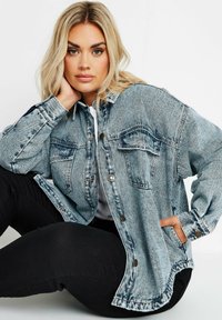 Ljusblå denimjacka med två bröstfickor, knappstängning och en avslappnad passform. Svarta byxor som är delvis synliga.