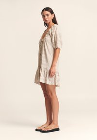 Gestreepte jurk met korte mouwen in beige met een gerimpelde zoom en strik aan de voorkant. Model draagt beige slippers met een casual ontwerp.