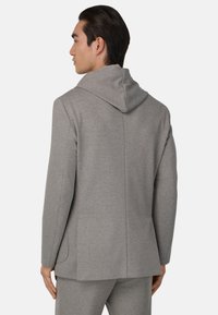 Blazer grigio in jersey con cappuccio, caratterizzato da un design snello, maniche lunghe e una fessura posteriore singola. Texture sottile e cuciture a vista.