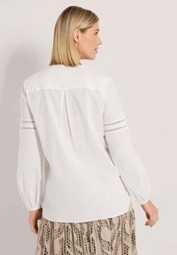 Frau mit kurzem blonden Haar, die eine weiße Bluse mit langen Ärmeln und Spitzenstreifen an den Ärmeln sowie einen beigen gemusterten Rock trägt, von hinten gezeigt.