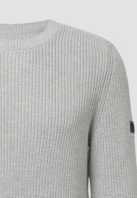 Grauer Strickpullover mit rundem Ausschnitt, strukturiertem Material, gerippten Bündchen und einem kleinen schwarzen Logopatch am linken Ärmel.