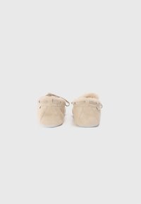 Chaussons en suède beige avec une douce doublure en fourrure, présentant un bout arrondi, des coutures minimales et un design à enfiler sans quincaillerie visible.