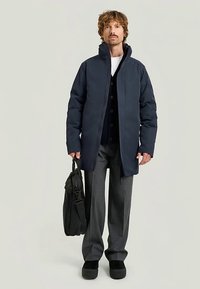 Mörkblå vadderad jacka med hög krage och dragkedja; bärs över en svart väst med knappar, grå byxor och svart väska. Svarta slip-on skor.