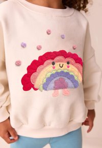Krémově zbarvená mikina s vyšitou barevnou duhou a usměvavou postavičkou, doplněná texturovanými pastelovými bambulkami.