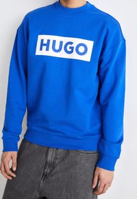 Niebieska bluza z okrągłym dekoltem i długimi rękawami, z dużym białym, prostokątnym logo z napisem "HUGO" na wysokości klatki piersiowej.