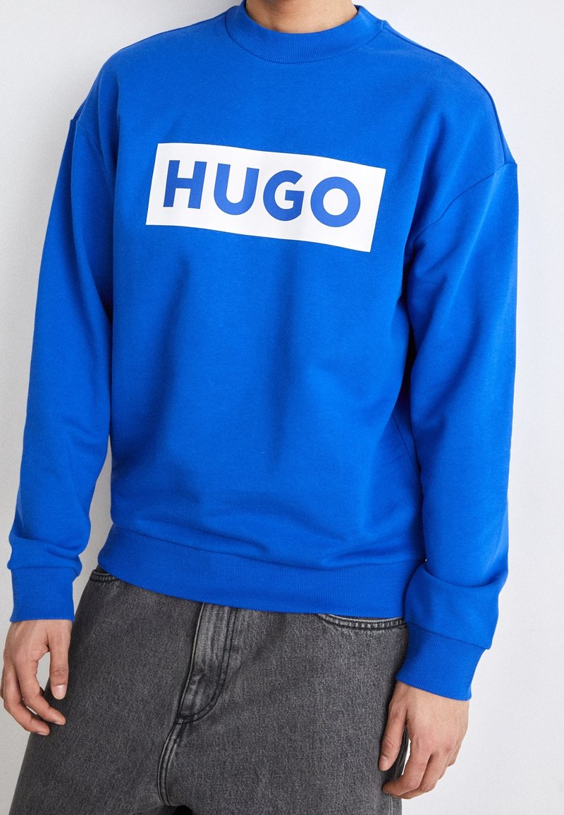 Niebieska bluza z okrągłym dekoltem i długimi rękawami, z dużym białym, prostokątnym logo z napisem "HUGO" na wysokości klatki piersiowej.