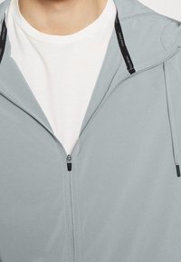 Světle šedá zipová bunda s hladkou texturou, s černým zipem a nastavovací šňůrkou, vrstvená přes bílou košili.