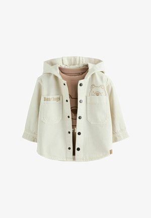 Hellcremefarbene Kapuzenjacke mit Frontknöpfen, zwei Brusttaschen, eine mit dem Text "Bear hugs", geschichtet über einem braunen Hemd mit Bärengesicht-Design.