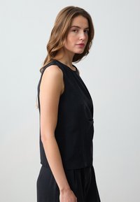 Top senza maniche nero caratterizzato da una texture liscia e opaca, scollatura rotonda e dettaglio drappeggiato sul davanti, progettato per un fit rilassato.