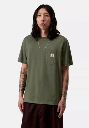 Hombre con cabello largo y oscuro que lleva una camiseta Carhartt verde oliva con bolsillo en el pecho, collar plateado y tatuajes en ambos antebrazos.