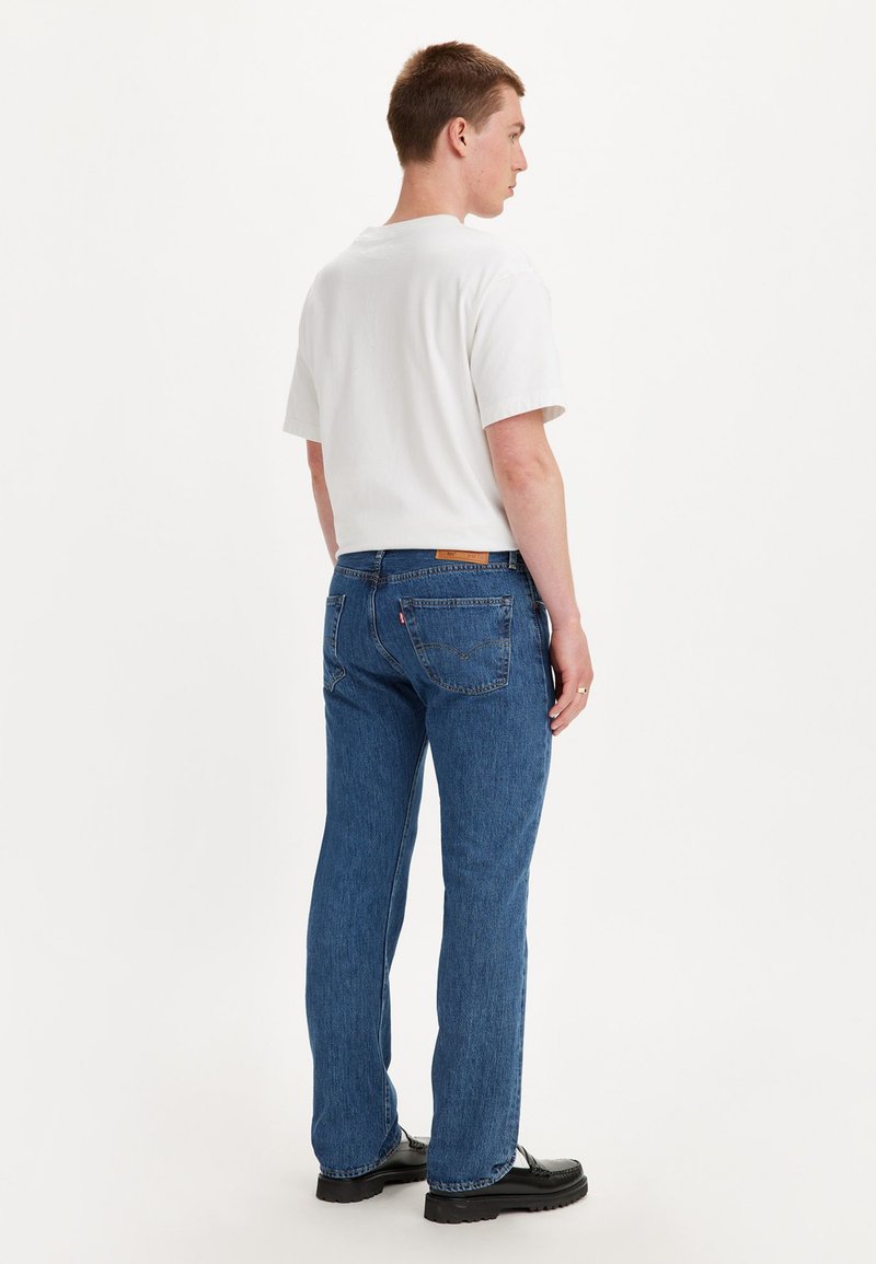 T-shirt blanche à manches courtes, jean en denim bleu à coupe slim, et chaussures noires. Le jean présente des poches arrière et une étiquette en cuir à la taille.