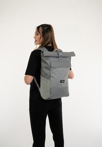 Sac à dos à rabat gris avec un panneau avant texturé, des bretelles réglables et une poche latérale. Il présente un patch logo sur le bas de l'avant.