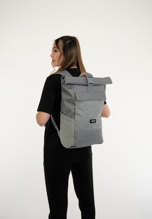 Zaino grigio a chiusura arrotolata con pannello frontale testurizzato, spallacci regolabili e una tasca laterale. Presenta un patch con logo sulla parte bassa anteriore.