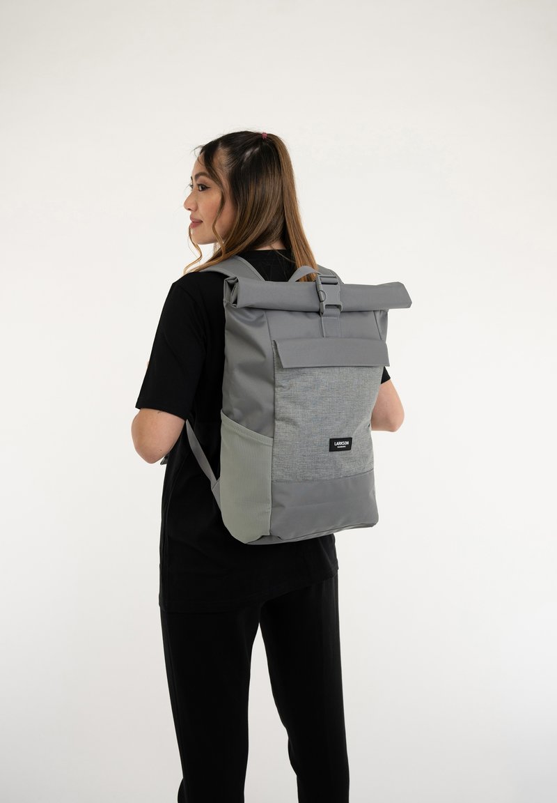 Sac à dos à rabat gris avec un panneau avant texturé, des bretelles réglables et une poche latérale. Il présente un patch logo sur le bas de l'avant.