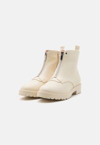 Botas de tobillo de color crema, hechas de cuero suave. Características incluyen un cierre de cremallera, punta redondeada y una suela gruesa y texturizada.