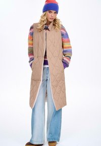 Beżowa pikowana długa kamizelka z kołnierzem i zapięciem na napy. Nałożona na sweter w wielobarwne paski, w połączeniu z jasnoniebieskimi spodniami o szerokich nogawkach.