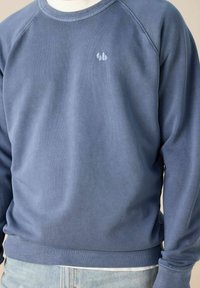 Homme portant un sweat-shirt bleu à col rond avec un petit logo brodé sur la poitrine et un jean en denim bleu clair.