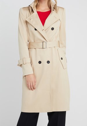 Beiger zweireihiger Trenchcoat mit schwarzen Knöpfen, tailliertem Gürtel und Ärmelschlaufen, getragen über einem roten Oberteil und schwarzen Hosen.