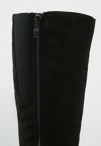 Botte en daim noir avec une fermeture éclair verticale sur le côté, dotée d'un panneau en textile élastique pour plus de flexibilité, d'une texture lisse et d'un design épuré.