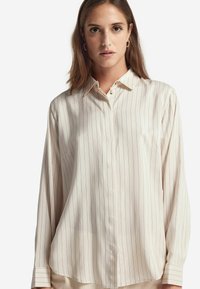 Camicia button-up color crema con un motivo a righe verticali, maniche lunghe, colletto strutturato e una texture di tessuto liscia.