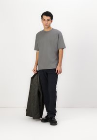 Šedé tričko s krátkým rukávem, tmavé vzorované kalhoty a zelená prošívaná bunda držena v ruce. Černé boty doplňují outfit.