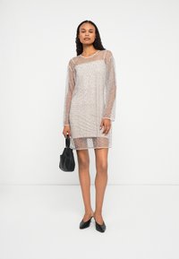 Egy áttetsző, hosszú ujjú, díszített ruhát viselő nő, alatta egy slip-el, kezében egy kis fekete táskával, és fekete, hegyes orrú cipőben.