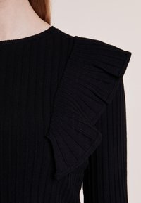Schwarzes, geripptes Stricktop mit einem Rüschendetail an einer Schulter, strukturiertem Stoff und klassischem Rundhalsausschnitt.