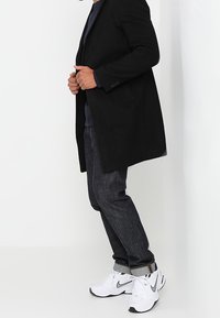 Zwarte wollen overjas, slim-fit donkere jeans en witte sportschoenen met zwarte accenten. De outfit heeft een gelaagde look met een casual stijl.
