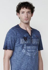 Sininen, lyhythihainen T-paita haalistuneella tekstuurilla, jossa on kolmesta napista koostuva henley-kaulus. Rintapielessä on painettu teksti ja koristeellinen kasvatus.