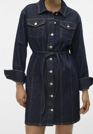Robe en jean bleu foncé boutonnée, avec poches à rabat devant, manches longues, ceinture nouée à la taille et détails de couture visibles.