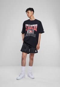 Juodas oversized t-shirt su raudona ir balta grafine tekstu, derinamas su juodais sportiniais šortais ir baltais sportbačiais. Švarus, urbanistinis stilius.