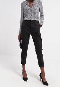 Vrouw in zwarte opgerolde broek, zwarte hoge hakken en een lange mouwen blouse met een patroon, met een zwarte clutch in één hand.