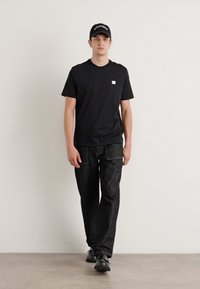 Zwarte korte mouw t-shirt met een klein logo, gecombineerd met donkerdenim jeans en zwarte schoenen, gedragen met een bijpassende pet, staand tegen een neutrale achtergrond.