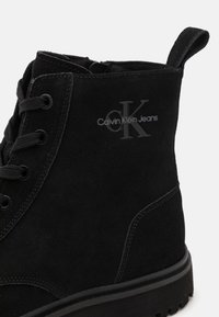 Stivale alla caviglia in suede nero con punta tonda, superficie testurizzata, stringhe frontali e logo Calvin Klein Jeans impresso sul lato. Suola in gomma.