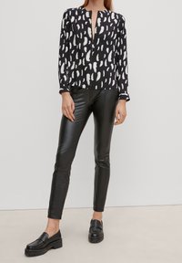 Blouse noire à manches longues avec un motif abstrait blanc, dotée d'une patte de boutonnage avant partielle, associée à un pantalon en faux cuir noir et des mocassins à plateforme.