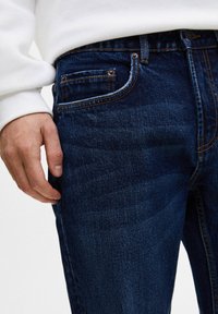 Jean en denim bleu foncé avec une texture lisse. Comprend cinq poches, une quincaillerie couleur cuivre et des coutures ton sur ton sur les coutures.