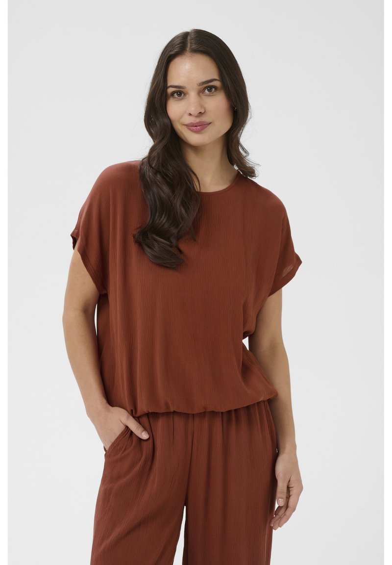 Femme aux longs cheveux foncés portant un top ample à manches courtes couleur rouille et un pantalon assorti, debout avec une main dans la poche.