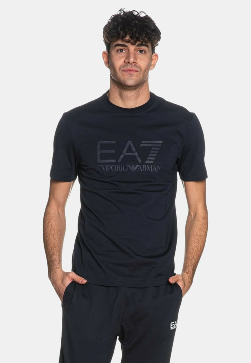 Mand med kort krøllet hår iført en sort EA7 Emporio Armani t-shirt og bukser, stående med hænderne i lommerne mod en hvid baggrund.