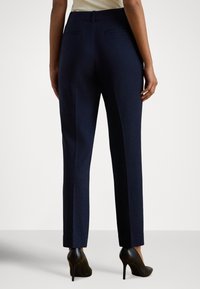 Calças de alfaiataria azul-marinho com corte slim e linhas limpas, com dois bolsos traseiros e textura de tecido suave. Usadas com sapatos de salto alto pretos.