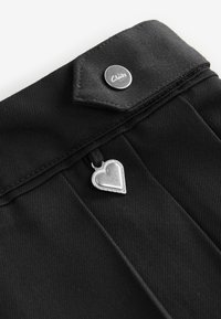 Sort stof med en metalknap og en sølvhjertevedhæng, der er fastgjort, med en tekstureret overflade og subtile plisseringer.