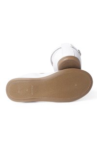 Weisse Schuhe mit einem glatten Lederobermaterial, verstellbarem Riemen und einer strukturierten braunen Gummisohle. Sie verfügen über eine runde Zehenform und ein minimales Design.