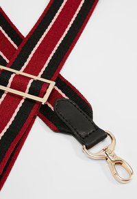 Punainen, musta ja vaaleanpunaraidallinen kangashihna, jossa kullanväriset metalliosat. Sisältää nahkaosan ja metallikiinnittimen kiinnitystä varten.