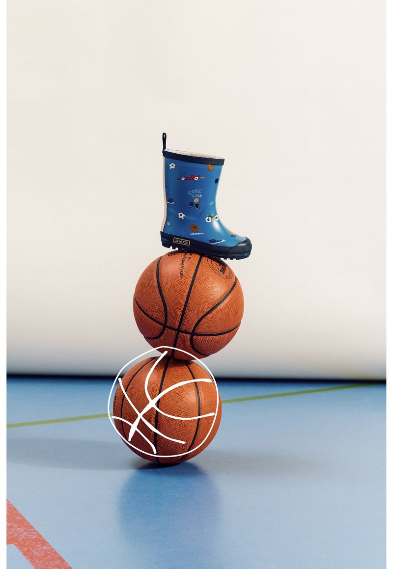 Une botte de pluie bleue pour enfants avec des motifs sportifs posée en équilibre sur deux ballons de basket empilés sur le sol d'un gymnase.