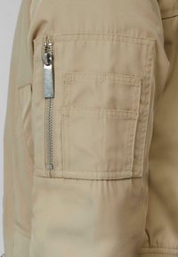Manche de veste bomber beige avec une fermeture éclair argentée et une poche à rabat, réalisée en tissu lisse avec des détails de couture visibles.