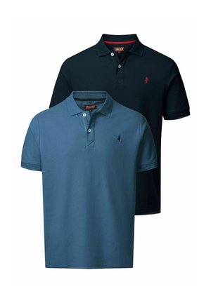 Man in een marineblauw poloshirt, blauwe spijkerbroek en bruine veterlaarzen die staat en naar beneden kijkt met een lichte glimlach.