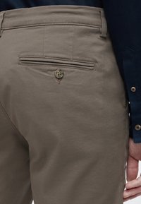 Κοντινό πλάνο πίσω τσέπης σε μπεζ παντελόνι με κούμπωμα κουμπιού, φορεμένο με πουκάμισο μακριάς μανίκας σκούρου μπλε, που δείχνει μανσέτα με κουμπί και χέρι.