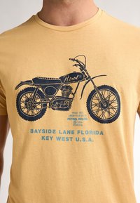 Žluté tričko s námořnickomodrým grafickým motivem vintage motocyklu, pod kterým je text "Bayside Lane Florida, Key West U.S.A."