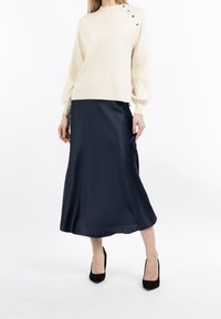 Pull-over crème à col rond avec trois boutons noirs en accent ; porté avec une jupe midi en satin marine et des escarpins noirs.