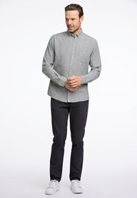Camicia grigia button-up con una trama sottile, taschino singolo sul petto, abbinata a pantaloni scuri e sneakers bianche, in piedi contro uno sfondo neutro.